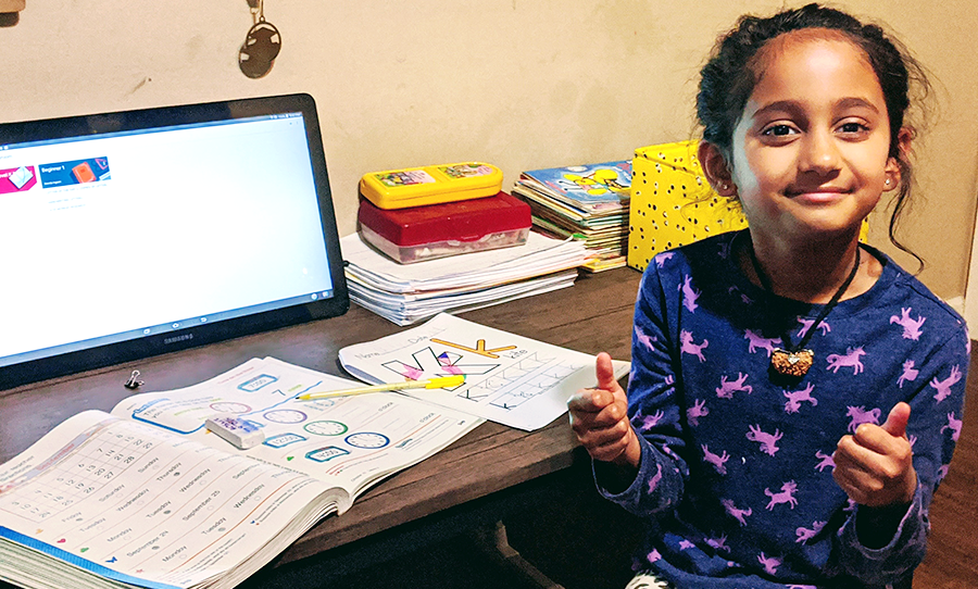 A Kindergarten student at her desk gives two thumbs up for at-home handwriting and learning.
