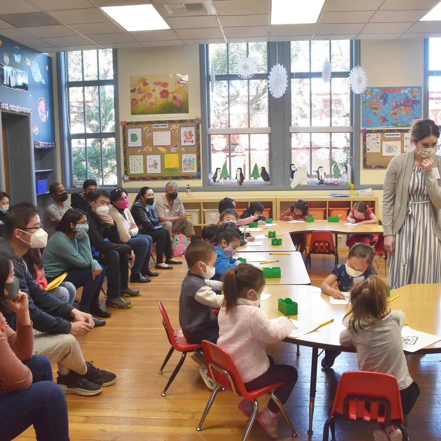 Image of a classroom with parents seated, observing in the back and children working diligently on alphabet pages at small tables. A female teacher looks at their work.